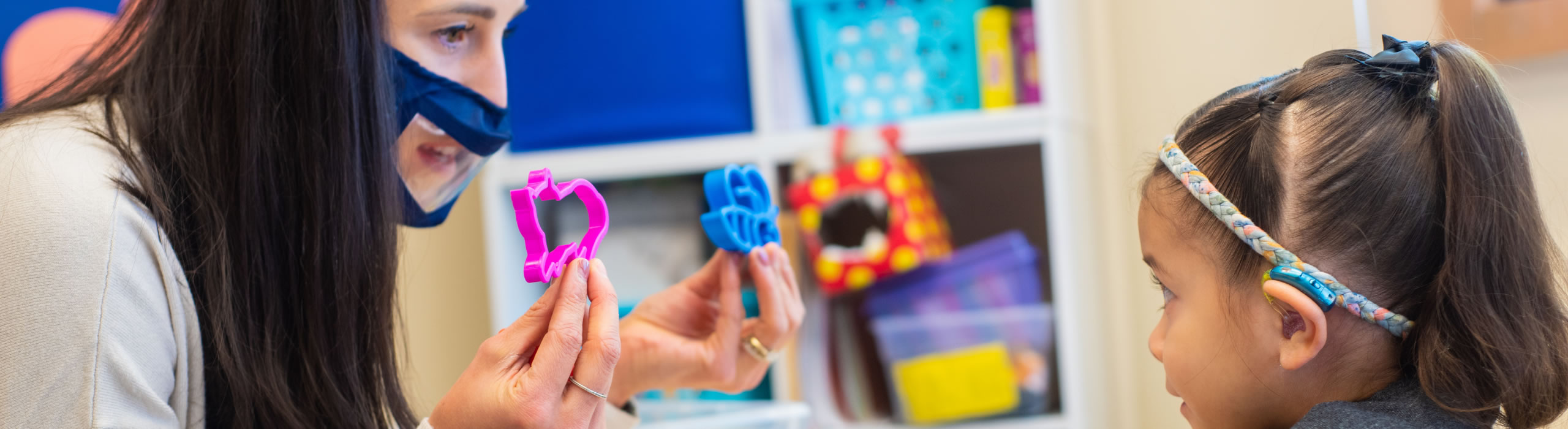Speech language pathologist working with young child to elicit language. Therapist is holding two objects- one pink and one blue – in each hand, and asking the child to choose one or the other, when prompted.